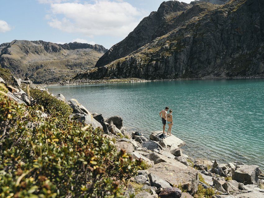 Informationen von A bis Z Zwei Personen am Ufer eines Bergsees mit klarem Wasser und felsiger Umgebung