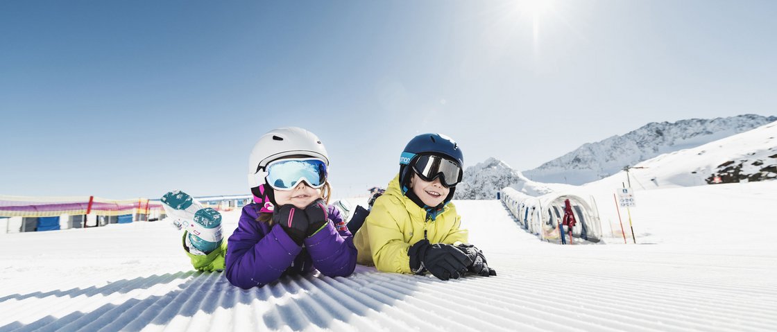 Your unforgettable ski holiday in Stubaital Two children lying on groomed snow in ski gear on a sunny mountain day