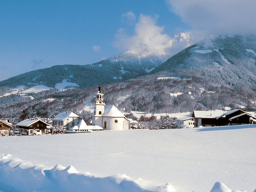Informationen von A bis Z Verschneites Dorf mit Kirche vor bewaldeten Bergen unter blauem Himmel