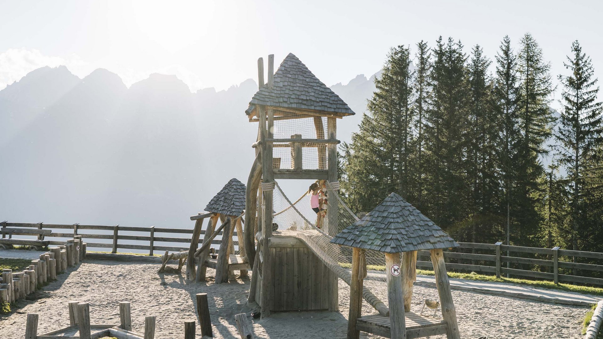 Highlights im Chalet in den Bergen Kinder spielen auf einem Holzspielplatz mit Netzbrücke in sonniger Berglandschaft