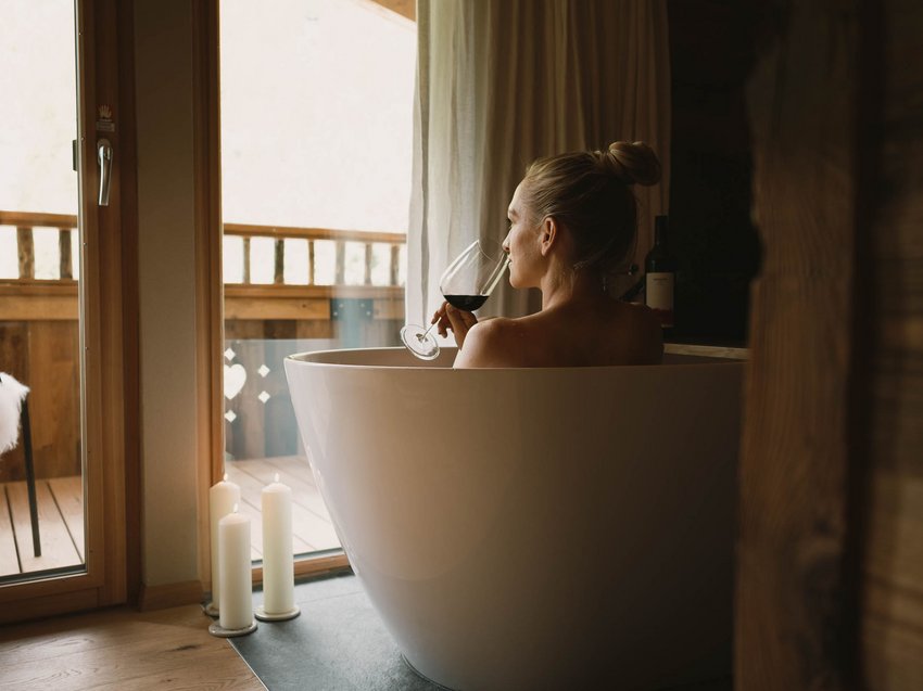 Ab ins Luxus-Chalet in Tirol Frau entspannt in Badewanne mit Rotweinglas neben Kerzen auf Holzboden