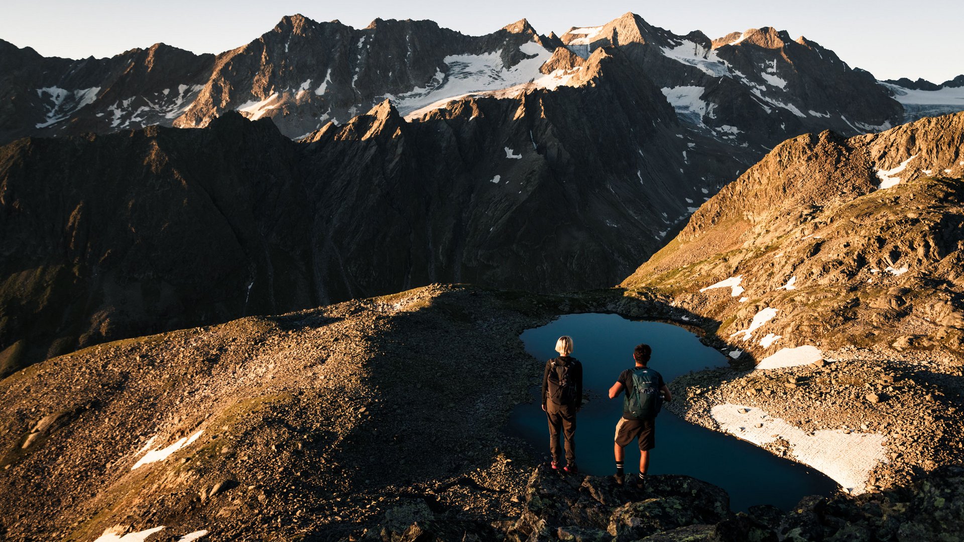 Zwei Wanderer stehen am Bergsee mit Blick auf schneebedeckte Alpenberge im Sonnenlicht